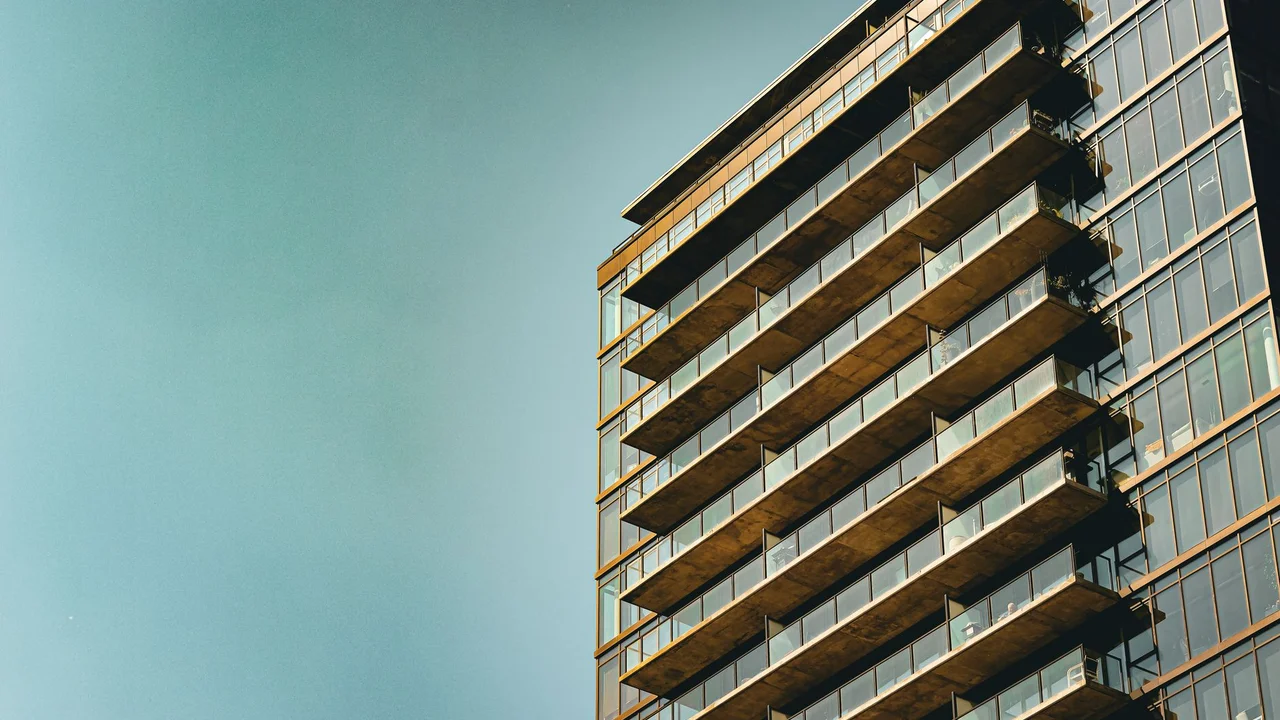 Sleek modern apartment building with glass facade and balconies, set against a clear blue sky.