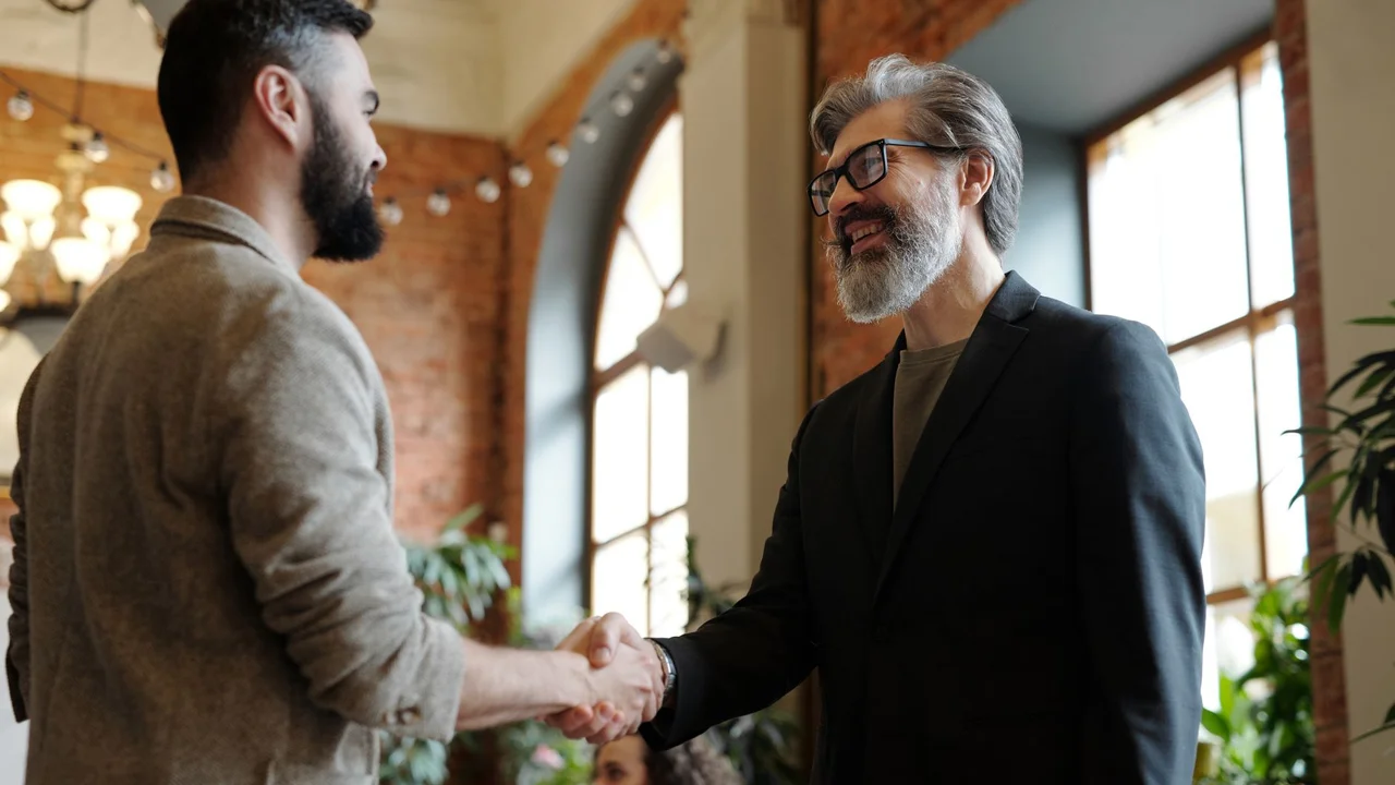 Two businessmen smiling during a handshake in a modern office setting, symbolizing success and partnership.
