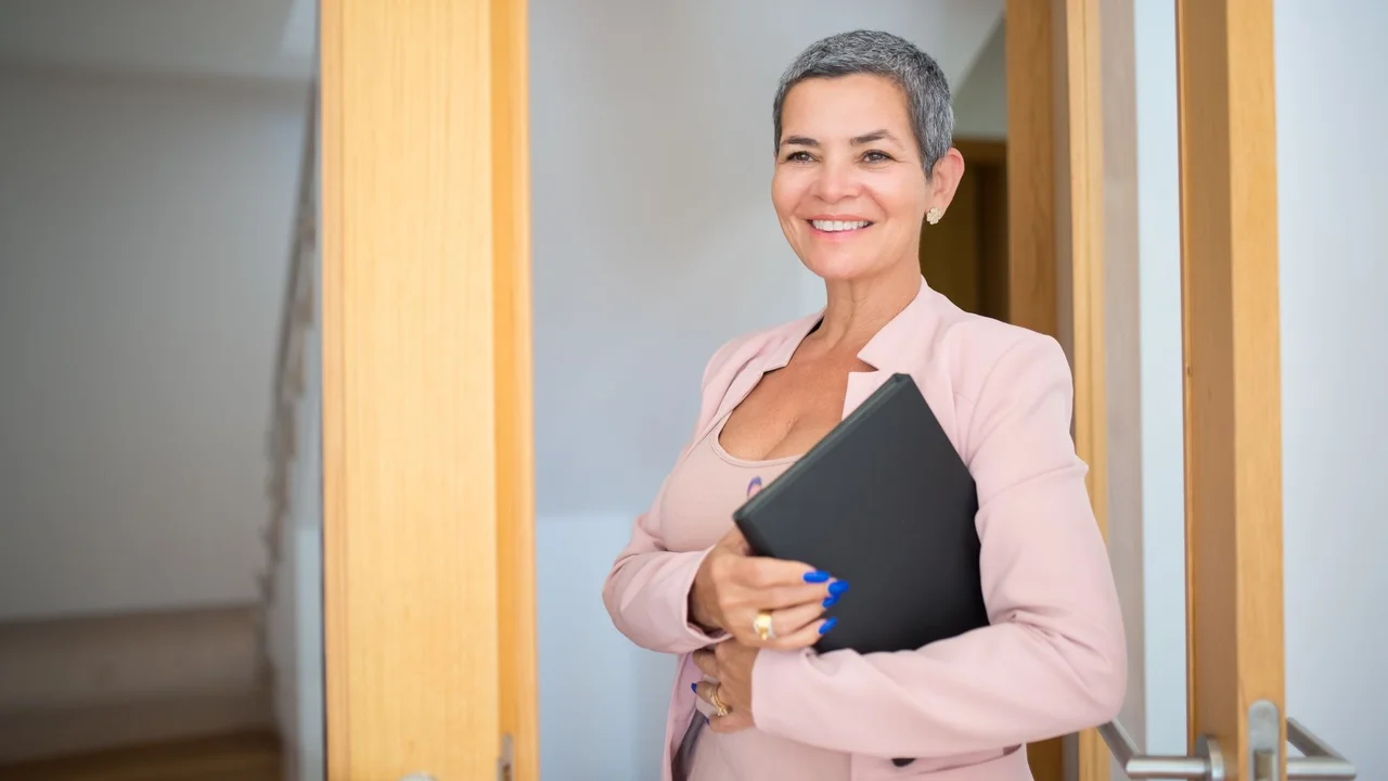 Smiling female real estate agent holding a black folder inside a modern building.